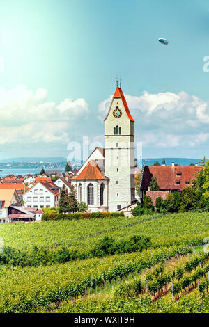 Panoramablick auf den Bodensee. Zeppelin, Apfelbäume und die Katholische Kirche St. Johann Baptist in Hagnau am Bild. Stockfoto