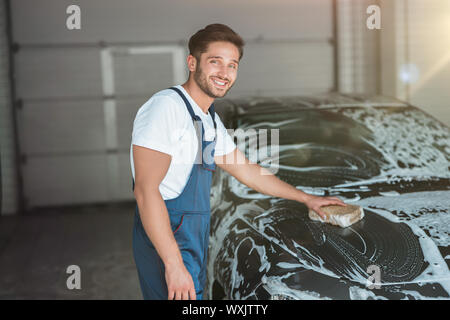 Jungen gutaussehenden Mann in Uniform waschen Auto mit Schwamm am Auto waschen entfernt. Stockfoto