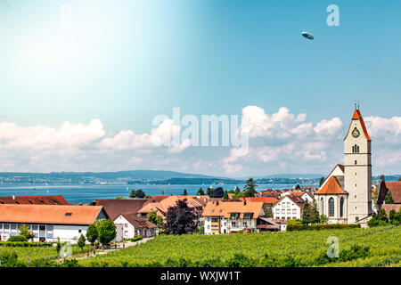 Panoramablick auf den Bodensee. Zeppelin und Katholische Kirche St. Johann Baptist in Hagnau am Bild. Stockfoto