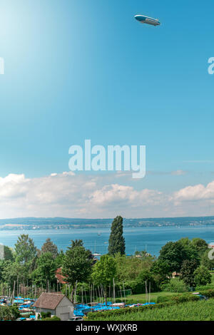 Panoramablick auf den Bodensee. Zeppelin, Apfelbäume und die Katholische Kirche St. Johann Baptist in Hagnau am Bild. Stockfoto
