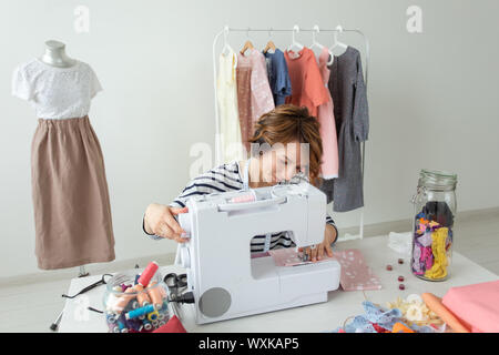 Junge talentierte Frau Designer Kleidung näht Ihr neues Produkt an Ihrem Schreibtisch sitzen mit Nähmaschine und Zubehör. Konzept der Hobbys und Berufe Stockfoto