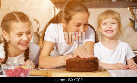 Family Portrait von Mutter und Töchter bereiten Geburtstag Kuchen zusammen, Mom ist Schneiden Biskuitteig mit Messer in der Küche. Stockfoto