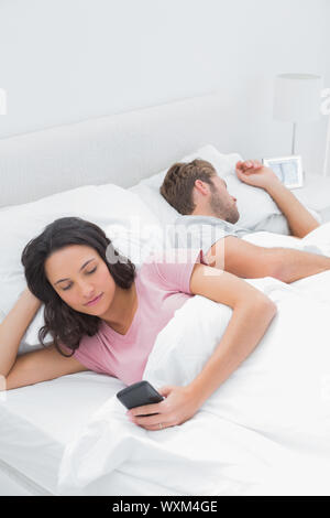 Frau mit ihrem Handy im Bett, während ihr Mann neben ihr schläft Stockfoto