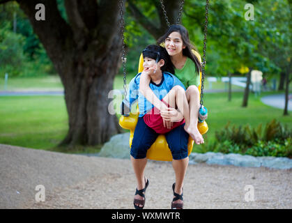 Große Schwester Betrieb deaktiviert Bruder auf speziellen braucht Schwung am Spielplatz im Park. Kind hat Zerebralparese. Stockfoto