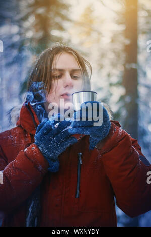 Der Kerl, der in einem roten warme Jacke und blaue Handschuhe fror während einer Wanderung durch den Winter dicken Kiefernwald, also das Trinken aus einer Tasse heißen Kaffee, während Smokin Stockfoto