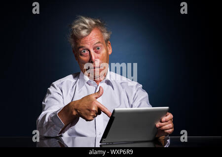 Emotionale Senior Business Mann mit ein ausdrucksstarkes Gesicht, der auf seinem Laptop in Horror und ungläubig, als er sitzt an einem Tisch gegen einen dunklen Studio hinterg Stockfoto