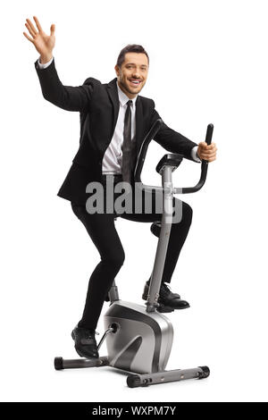 Volle Länge Schuß eines lächelnden jungen Geschäftsmann Training auf einem stationären Fahrrad und winkend auf weißem Hintergrund Stockfoto