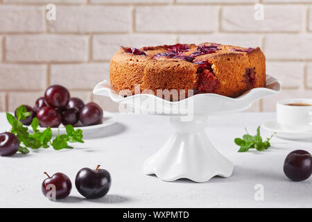 Pflaume Torte auf einem weißen Kuchen stand mit frischen Pflaumen auf Tisch und Wand im Hintergrund, close-up Stockfoto