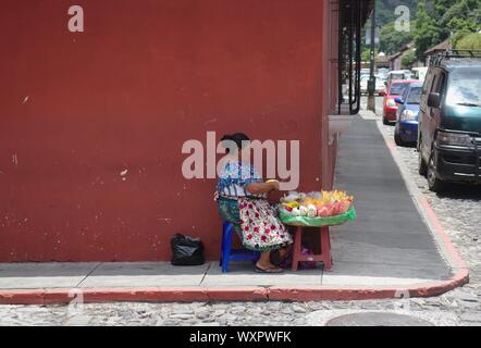 Antigua, Guatemala - August 2019: Weibliche Gemüse Anbieter sitzen auf Stuhl aus Kunststoff auf dem Bürgersteig im Schatten während der Zubereitung von Mahlzeiten. Stockfoto