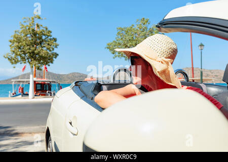 Single glücklich reife Frau mit Hut und Sonnenbrille fahren Ihr Verdeck Automobil auf hellen, sonnigen Tag in der Nähe von Ocean. Die Frau blickt zurück in Richtung Stockfoto