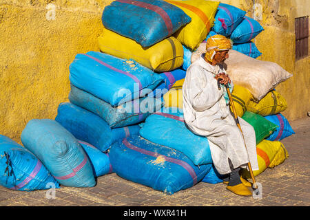 Ältere Menschen traditionell gekleidete Marokkaner mit Stock sitzen auf bunten Kunststoff Ballen von Lebensmittelzutaten, die von der gelben wand, Marrakesch, Marokko Stockfoto