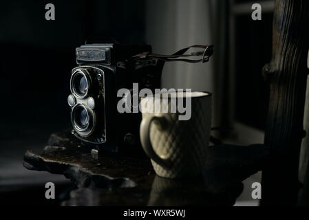 Foto mit dunklem Motiv einer klassischen Kamera entlang einer Tasse auf einer Ebenen Oberfläche Stockfoto
