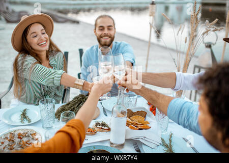 Gruppe von jungen Freunden Spaß haben, klirren Gläser während einer festlichen Abendessen in der wunderschön dekorierten Tisch in der Nähe der See im Freien Stockfoto