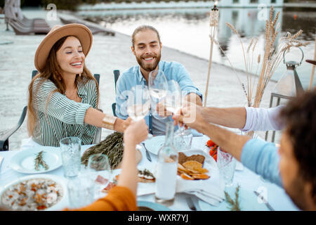 Gruppe von jungen Freunden Spaß haben, klirren Gläser während einer festlichen Abendessen in der wunderschön dekorierten Tisch in der Nähe der See im Freien Stockfoto