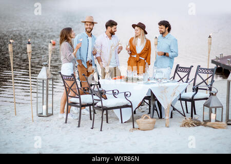 Junge Freunde heraus zusammen hängen, stehend mit Weingläsern in der Nähe der wunderschön dekorierten Esstisch am Strand bei Sonnenuntergang Stockfoto
