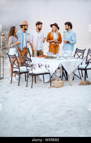 Junge Freunde heraus zusammen hängen, stehend mit Weingläsern in der Nähe der wunderschön dekorierten Esstisch am Strand bei Sonnenuntergang Stockfoto