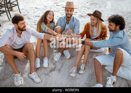 Gruppe junger Freunde klirren Gläser während einer festlichen Sitzung, Genießen am Abend am Sandstrand Stockfoto