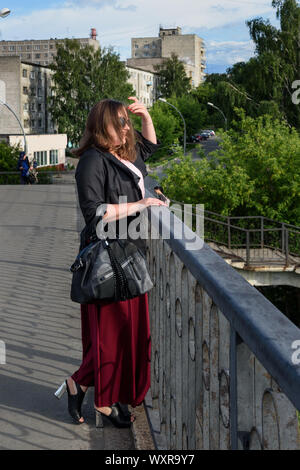 Brunette Mädchen auf der Brücke in der Stadt. Mädchen in schwarzen Jacke und roter Rock mit langen Haaren im Sommer Tag. Plus size Model Stockfoto