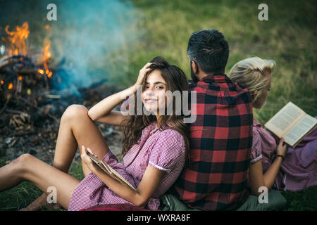 Camping in der Wildnis. Drehen Sie wieder Kerl auf der Suche nach Feuer, während zwei hübsche Mädchen Buch lesen. Smiling brunette mit klammern sich an ihre Freundin. Stockfoto