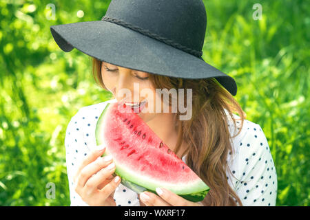 Hübsche junge Frau ist Wassermelone in Hut auf grüne Natur Hintergrund Essen, erwachsene Mädchen isst ein Stück Wassermelone outdoor Stockfoto