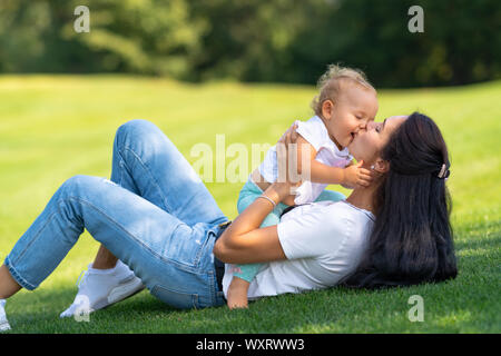 Liebevolle junge Mutter ihre kleine Tochter zärtlich küssen, als sie zusammen auf grünem Gras draußen im Garten im Schatten eines Baumes spielen Stockfoto