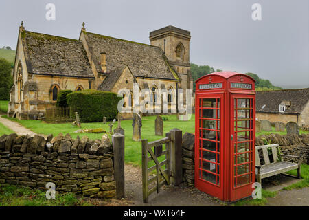 Rote Telefonzelle im 15. Jahrhundert der hl. Barnabas anglikanische Kirche mit Friedhof und Mauer Tor in feuchten regnerischen Wetter in Snowshill Cotswold England Stockfoto