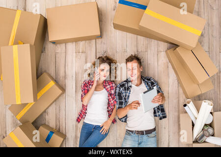 Umzug. Junger Mann browsing digitalen tablet, während Frau Reden auf dem Smartphone freudigen Lächeln lag auf dem Boden unter den Boxen Ansicht von oben Stockfoto