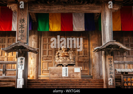 DEC 3, 2018 Yamagata, Japan - Japanische geschnitztem Holz lächelnden Buddha Statue mit alten Holz Wand und bunte Fahne von Konponchudo Haupthalle im yamadera Ri Stockfoto