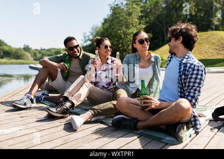 Freizeit-, Picknick- und Personen Konzept - Freunde, trinken Bier und Apfelwein vom See Pier im Sommer Park Stockfoto
