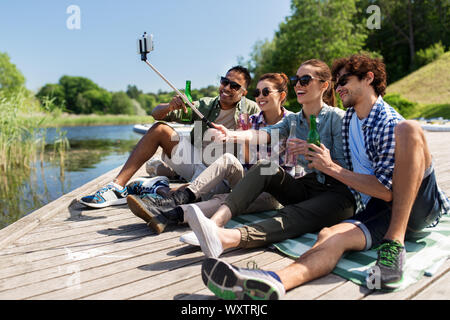 Freizeit-, Picknick- und Technologiekonzept - Freunde mit Getränken, Bild von selfie Stick auf See Pier im Sommer Park Stockfoto