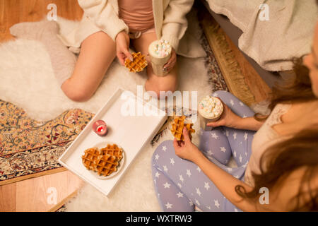 Freundschaft, hygge und pyjamaparty Konzept - zwei weibliche Freunde oder Teenager trinken heiße Schokolade mit Marshmallows und essen Waffeln zu Hause Stockfoto