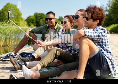 Freizeit-, Picknick- und Technologiekonzept - Freunde mit Getränken, Bild von selfie Stick auf See Pier im Sommer Park Stockfoto