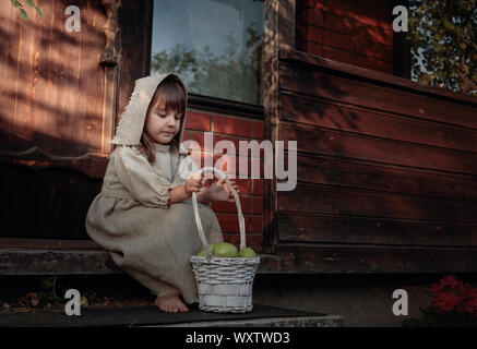 Kleine barfüßige Mädchen in einem alten Leinwand Kleid und eine Kappe auf die Schwelle des Hauses. Mädchen mit Äpfeln an einem Sommerabend in der Nähe des alten Hauses. Stockfoto