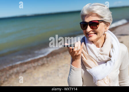 Alte Menschen und Freizeitaktivitäten Konzept - Glücklich lächelnde ältere Frau mit Voice Command Recorder auf dem Smartphone, am Strand in Estland Stockfoto