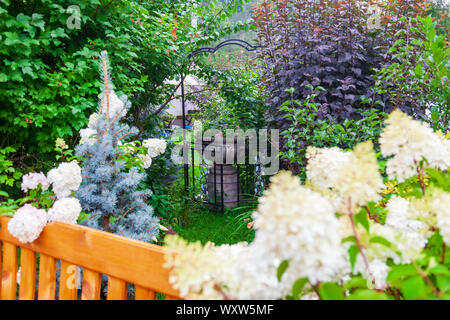 Blühender Garten im Innenhof eines Landhauses mit einer Vase und einem Bett aus Stein mit Blumen von grünen Pflanzen und Bäumen umgeben. Stockfoto