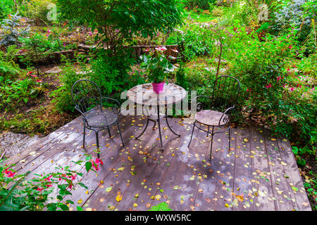 Blühender Garten im Innenhof eines Landhauses mit zwei Stühlen und Tisch von grünen Bäumen und Blättern umgeben. Rest und Picknick in der Natur. Stockfoto