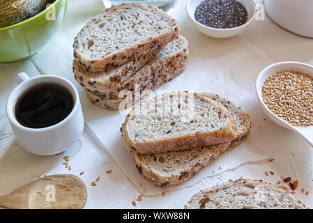 Braun Brot mit Getreide neben einer Tasse Kaffee auf einem weißen Tisch von einem rustikalen Küche. Samen und Zutaten für zu Hause Bäckerei. Stockfoto