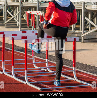 Ein High School Mädchen Leichtathlet ist in der Aufwärmphase für ihre Hurdle Race zu Fuß über Hürden auf einer roten Piste in Ihrem sweatsuit. Stockfoto