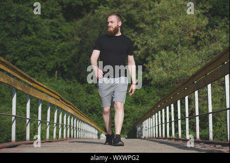 Aktiven jungen erwachsenen Mann mit Bart in schwarzen T-Shirt und Sneakers entlang hölzerne Brücke in der natürlichen Parklandschaft während cardio Workout Stockfoto