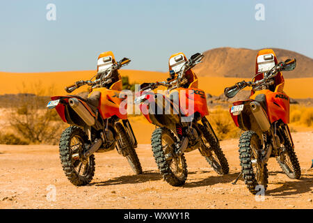 Merzouga, Marokko - Februar 24, 2016: 3 orange Motorräder aufgereiht bereit auf einer Wüste Sahara Abenteuer zu gehen. Sie sitzen unter einem strahlend blauen Himmel auf Stockfoto