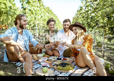 Junge Freunde bei einem Picknick mit vielen leckeren Speisen und Wein, zusammen zu sitzen und Spaß haben auf der Picknickdecke im Weinberg Stockfoto