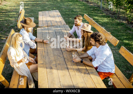 Eine Gruppe von jungen Menschen, Wein trinken und zusammen reden, während am Esstisch im Freien sitzen auf dem Weinberg an einem sonnigen Abend Stockfoto