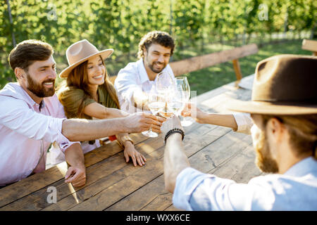 Eine Gruppe von jungen Menschen, Wein trinken und zusammen reden, während am Esstisch im Freien sitzen auf dem Weinberg an einem sonnigen Abend Stockfoto