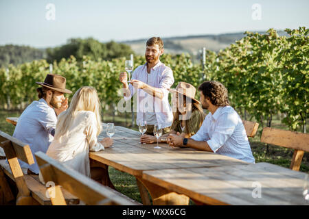 Eine Gruppe von jungen Menschen, Wein trinken und zusammen reden, während am Esstisch im Freien sitzen auf dem Weinberg an einem sonnigen Abend Stockfoto