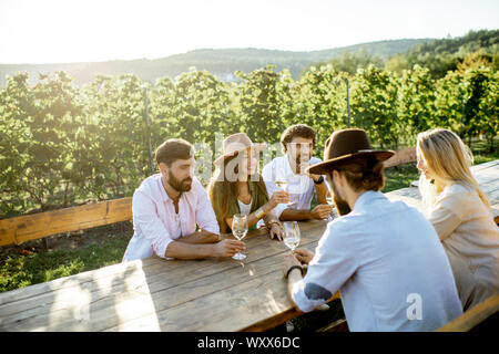 Eine Gruppe von jungen Menschen, Wein trinken und zusammen reden, während am Esstisch im Freien sitzen auf dem Weinberg an einem sonnigen Abend Stockfoto