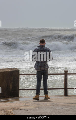 Ein junger Mann stand auf einer Wand mit Blick auf das Meer an einem stürmischen Meer an einem windigen Tag an der Küste. Stockfoto