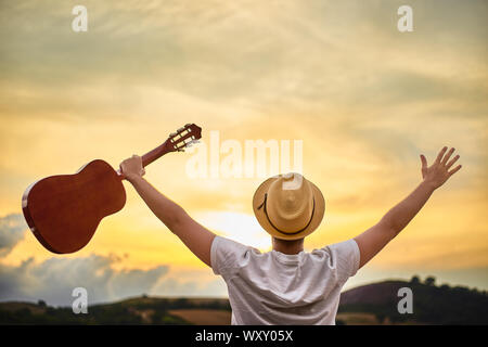 Ein junger kaukasischer Mann wandte sich mit einem Strohhut und ein weißes T-Shirt, grüßt die untergehende Sonne über den Hügeln mit offenen Armen und hält einen klassischen Stockfoto