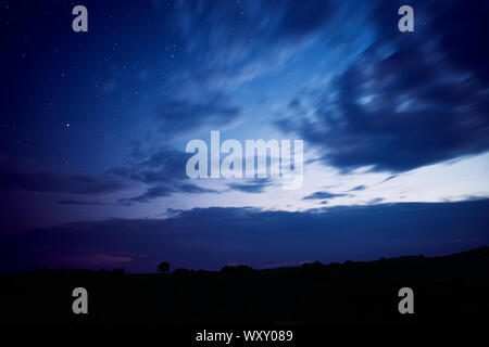 Nacht Landschaft und während der Blauen Stunde, mit der Silhouette eines einsamen Baum unter den Hügeln, den Sternenhimmel und voller Wolken, Langzeitbelichtung Stockfoto