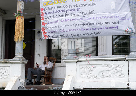 Ein Mann sitzt auf der Veranda in einem Schaukelstuhl, raucht und schaut auf sein Handy. Ein thunfischwadenfänger Veranda hängt ein riesiges Transparent mit den Sieb Stockfoto