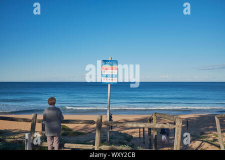 Eine Frau neben einem hölzernen Zaun am Eingang zum Palm Beach mit einem Warnschild an einem sonnigen Tag mit einem klaren blauen Himmel, New South Wales, Australien Stockfoto